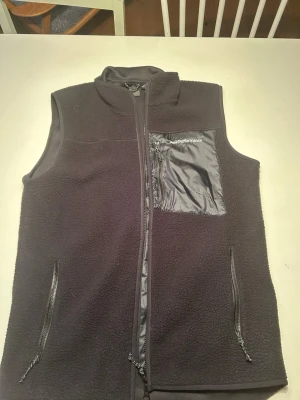 Svart fleeceväst - Peak Performance  - Mysig svart fleeceväst från Peak Performance med krage, hel dragkedja framtill och tre fickor med dragkedja, varav en bröstficka i blankt material. Samt 2st innerfickor. Perfekt till lite kyligare kvällar samt funkar den till en stiligare outfit. 