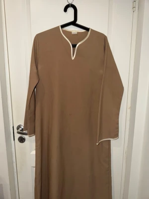 Abaya Beige långklänning med vit kant - En enkel och stilren beige långklänning med vita kanter vid halsringning och ärmslut. Klänningen har lång ärm och rak passform, perfekt för en avslappnad och bekväm look.