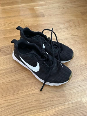 Svarta Nike sneakers med vit sula - Svarta sneakers från Nike med vit sula och stor vit swoosh-logga på sidan. Ovandelen är i meshmaterial för bra andningsförmåga och skorna har klassisk snörning. Perfekta för en sportig och avslappnad stil.