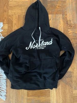 Svart hoodie med Norrland-tryck - Svart hoodie med vit text 'Norrland' på bröstet och justerbar huva med vita snören. Klassisk känguruficka framtill och avslappnad passform. Perfekt för dig som gillar streetwear och vill ha något bekvämt och stilrent.