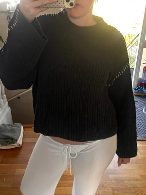 Svart ribbad stickad tröja - Mysig svart stickad tröja med ribbad struktur och vita dekorativa sömmar längs ärmar och axlar. Tröjan har en croppad passform och långa ärmar, perfekt för lager på lager under kyligare dagar.
