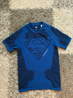 YoungLA superman compression - Blå träningströja från YoungLA med stort Superman-tryck på bröstet. Tröjan har korta ärmar och coola mönstrade paneler på sidorna och ärmarna. Perfekt för gymmet eller sportaktiviteter. Materialet är syntet och andas bra. Oanvänd, kvitto finns.