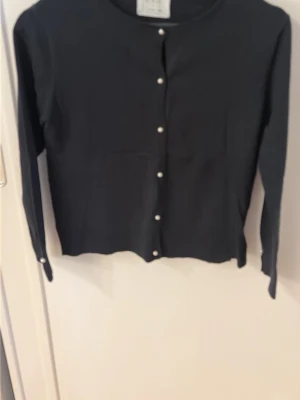 Svart kofta med pärlknappar från ZARA - Stilren svart kofta från ZARA med långa ärmar och pärlknappar framtill. Klassisk rund halsringning och rak passform. Perfekt att matcha med jeans eller kjol för en enkel men snygg look. Storlek 152cm. 11-22 år. 