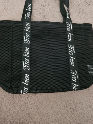 Svart mesh shoppingväska Tres bien - Snygg svart shoppingväska i meshmaterial med breda axelband där det står 'Tres bien' i vit text. Väskan har en rektangulär form och är lätt och luftig, perfekt för att bära med sig till stranden eller stan. Enkel men trendig design som sticker ut.