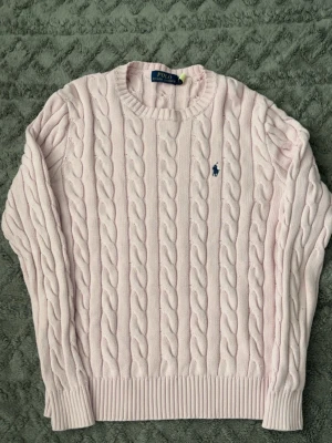 Ljusrosa kabelstickad tröja Polo Ralph Lauren - Supersnygg ljusrosa kabelstickad tröja från Polo Ralph Lauren. Klassisk rund halsringning, ribbade muddar och ikonisk blå logga broderad på bröstet. Perfekt för dig som gillar en clean och tidlös look.
