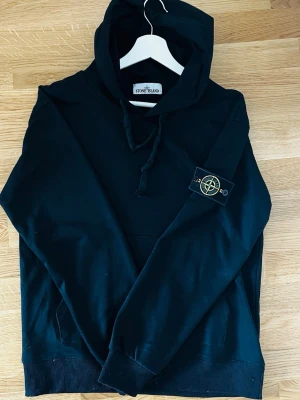 Svart hoodie från Stone Island - Svart hoodie från Stone Island med klassisk huva och dragsko. Har en stor ficka framtill och den ikoniska Stone Island-loggan på vänster ärm. Perfekt för en avslappnad och stilren look. Tillverkad i mjukt material för extra komfort.
