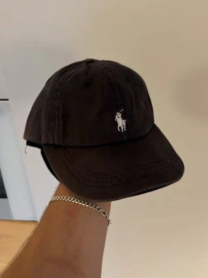 Ralph Lauren keps - Svart keps från Polo Ralph Lauren med klassisk böjd skärm och broderad vit logga framtill och baktill. Justerbar rem baktill för perfekt passform. Stilren och enkel design som passar till många outfits.