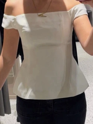 Vit offshoulder topp med utsvängd midja - Stilren vit topp med offshoulder-design och utsvängd midja. Toppen är axelbandslös och har en elegant, figurnära passform som ger en snygg siluett. Perfekt för dig som gillar en clean och modern look.
