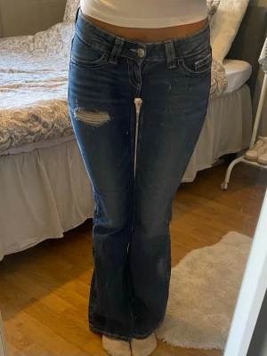 Mörkblå bootcut jeans med brodyr - Snygga mörkblå bootcut jeans med slitning på ena låret och broderade detaljer på bakfickorna. Jeansen har klassisk femficksdesign och låg midja. Perfekta för dig som gillar en avslappnad men trendig stil.