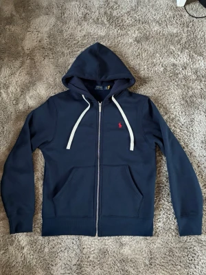 Mörkblå hoodie från Polo Ralph Lauren - Snygg mörkblå hoodie med dragkedja från Polo Ralph Lauren. Den har vit dragsko, huva och två stora fickor framtill. Ikonisk röd logga broderad på bröstet. Materialet är mjukt och bekvämt, perfekt för chill dagar.