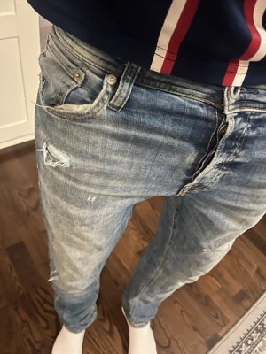 Ljusblå slitna jeans herr - Säljer ett par ljusblå jeans med slitningar och hål på vänster lår. Jeansen har klassisk femficksmodell, normal midja och smal passform. Perfekta för en avslappnad och trendig stil. Materialet är denim i bomull.