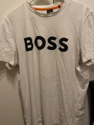 Vit BOSS t-shirt med tryck - Vit t-shirt från BOSS med stort svart BOSS-tryck på bröstet. Klassisk rund halsringning och korta ärmar. Tillverkad i mjuk bomull för en bekväm känsla. Perfekt för en clean och stilren look.