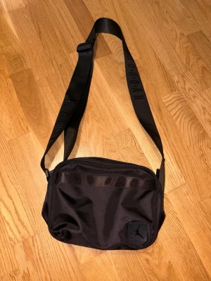 Svart axelväska från Jordan - Snygg svart axelväska från Jordan med justerbar axelrem och diskret logga framtill. Väskan har en rektangulär form och är tillverkad i slitstarkt syntetmaterial. Perfekt för att bära det viktigaste och ger en sportig vibe till din outfit.