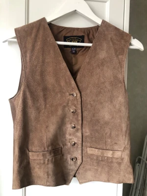 Beige mockaväst från H&M - Snygg beige mockaväst från H&M med klassisk V-ringning och knappar framtill. Västen har två fickor och är gjord i mjukt mockamaterial som ger en cool vintagekänsla. Perfekt att styla över en skjorta eller t-shirt.