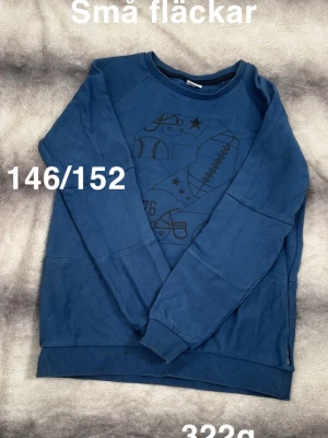 Blå sweatshirt  - Blå sweatshirt med tryck av olika sportprylar på bröstet, som baseboll, amerikansk fotboll och hjälm. Tröjan har ribbade muddar vid ärmslut, hals och nederkant. Perfekt för dig som gillar sportig stil.🌸 Små fläckar. Storlek 146/152