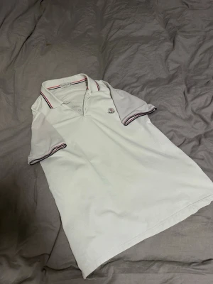 Vit Moncler piké - Vit pikétröja från Moncler med klassisk krage och knappar framtill. Tröjan har röda och blå ränder på krage och ärmslut samt Moncler-logga på bröstet. Storlek XL, passar som L/M. Svaga fläckar under armhålorna tyvärr. Annars i rätt så fint skick! Hör av er vid frågor/prisförslag! ✌️