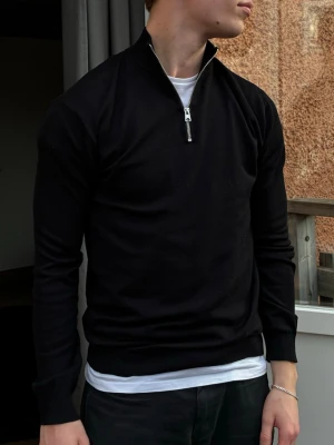 Svart stickad tröja med halfzip  - Svart stickad tröja med hög krage och halv dragkedja framtill. Tröjan har lång ärm, ribbade muddar och en slim passform. Perfekt att bära över en t-shirt för en clean look. Materialet ser ut att vara mjukt och följsamt.