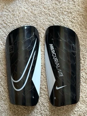 Nike Mercurial Lite benskydd svart/vit - Säljer ett par svarta Nike Mercurial Lite benskydd med vit detalj och stor Swoosh-logga. Lätta och smidiga, perfekta för fotbollsträning eller match. Snygg design med textur och tydlig märkning för vänster och höger ben.