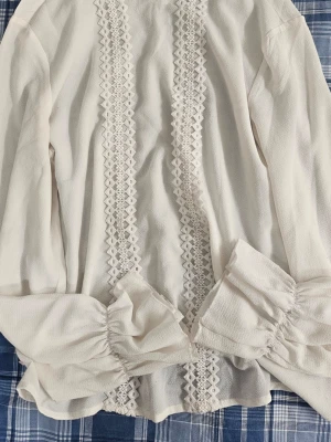 Vit blus med spetsdetaljer - Supersöt vit blus med långa ärmar och breda muddar. Framsidan har två rader dekorativ spets som ger en romantisk känsla. Blusen är gjord i ett lätt, strukturerat tyg som känns luftigt och bekvämt.