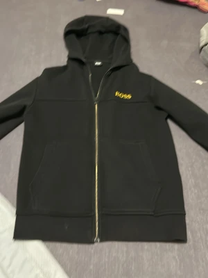 Svart hoodie med dragkedja från BOSS - Snygg svart hoodie från BOSS med guldfärgad logga på bröstet och dragkedja framtill. Klassisk huva och två stora fickor framtill. Tillverkad i mjukt material som känns skönt mot huden. Perfekt för en chill och stilren look.