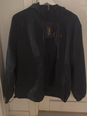 Mörk blå vindjacka från Polo Ralph Lauren - Snygg svart vindjacka från Polo Ralph Lauren med huva och dragkedja framtill. Jackan har en liten röd logga broderad på bröstet och justerbar nederkant med dragsko. Tillverkad i lätt och tåligt material, perfekt för blåsiga dagar.