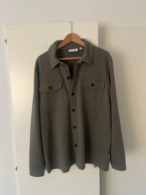 Olivgrön overshirt från Only & Sons - Stilren olivgrön overshirt från Weekday med klassisk krage, två bröstfickor med lock och knappar framtill. Overshirten har raka ärmar med manschett och är tillverkad i ett mjukt, slätt material. Perfekt för lager-på-lager-stil. Storlek XL och knappt använd. Jag säljer den för att den är för stor och har inte kommit till användning. Hör av er vid frågor!