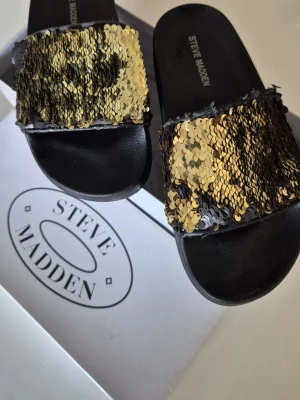 Steve Madden sandaler med paljetter - Svarta sandaler från Steve Madden med bred rem täckt av vändbara paljetter i guld och svart. Snygg slip-in modell med mjuk sula, perfekt för att sticka ut. Paljetterna kan ändra färg beroende på hur de dras, vilket ger en cool effekt. Med box.
