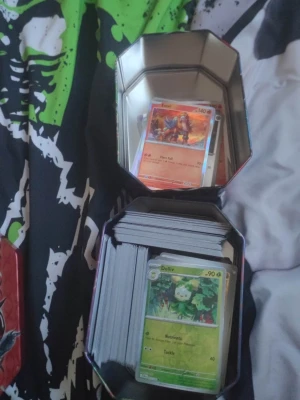 Stor samling Pokémonkort i plåtlåda - Säljer en stor samling Pokémonkort i en fyrkantig plåtlåda. Blandade kort med olika motiv, färger och energityper, inklusive holografiska och vanliga kort. Perfekt för dig som vill utöka din samling eller börja spela Pokémon TCG.