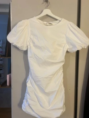 Vit kort puffärmad klänning med knyt - Trendig vit kort klänning med puffärmar och rund hals. Klänningen har öppen rygg med dubbla knytband och lätt rynkade detaljer längs sidorna. Perfekt för dig som vill ha en söt och modern look.