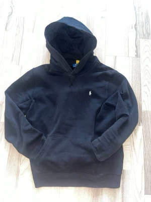 Svart hoodie från Polo Ralph Lauren - Klassisk svart hoodie från Polo Ralph Lauren med broderad logga på bröstet. Hoodien har en stor huva och magficka framtill. Tillverkad i mjukt bomullsmaterial som känns skönt mot huden. Perfekt för en avslappnad och stilren look.