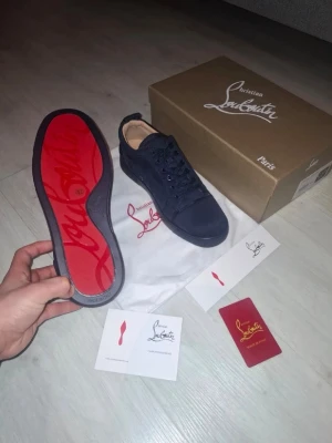 (ÄKTA) Christian Louboutin skor - Snygga svarta sneakers från Christian Louboutin med klassisk röd sula och diskret logga. Skorna har lågt skaft, svarta skosnören och är tillverkade i mocka. Perfekta för dig som vill ha en exklusiv och stilren look.