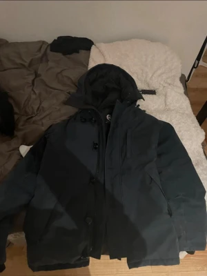 Svart parkas från Canada Goose - Säljer en mörkblå parkas från Canada Goose med klassisk patch på ärmen och huva. Jackan har dragkedja och knappar framtill samt flera fickor. Perfekt för kalla dagar och riktigt snygg streetstil.