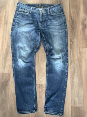 Blå jeans från Lee med slitningar - Säljer ett par klassiska blå jeans från Lee med snygga slitningar och tvättade detaljer. Jeansen har fem fickor, raka ben och normal midja. Materialet är denim i bomull och passformen är avslappnad men ändå stilren.