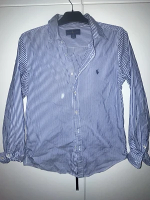 Blårandig skjorta från Ralph Lauren - Klassisk blårandig skjorta från Ralph Lauren med vit knappstängning och lång ärm. Skjortan har en liten broderad logga på bröstet och är tillverkad i mjuk bomull. Perfekt för en clean och stilren look.