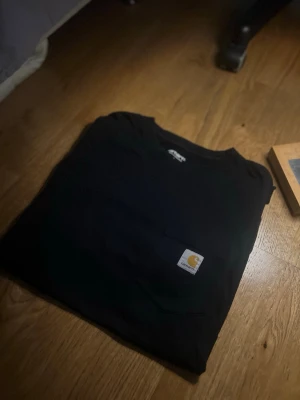 Svart Carhartt t-shirt med ficka - En svart t-shirt från Carhartt med klassisk rund hals och en praktisk bröstficka med Carhartt-loggan. T-shirten har korta ärmar och är tillverkad i mjuk bomull, perfekt för en avslappnad och stilren look.