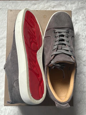 Gråa mocka skor med röd sula - Snygga gråa sneakers i mjuk mocka med klassisk röd sula och vita kanter. Skorna har gråa skosnören, diskreta sömmar och beige innersula i läder. Perfekta för dig som vill ha något exklusivt och stilrent. Skick 9/10 Inga defekter