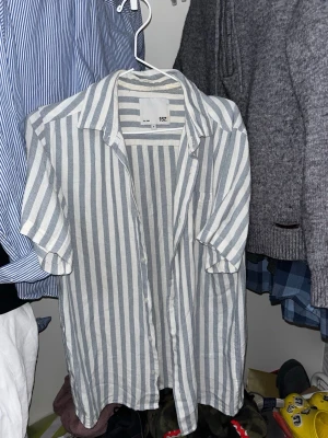 Randig skjorta från 157, kort ärm - Säljer en snygg skjorta från 157 med vertikala grå och vita ränder. Skjortan har korta ärmar, klassisk krage och knäppning framtill. Tillverkad i ett lätt och luftigt material, perfekt för varmare dagar.