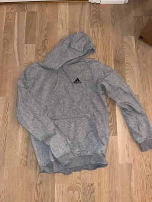 Grå hoodie från Adidas - Snygg grå hoodie från Adidas med klassisk logga på bröstet. Hoodien har huva med dragsko, magficka och ribbade muddar vid ärmar och nederkant. Tillverkad i mjukt bomullsmaterial som är skönt mot huden.