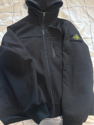 Svart vindjacka från Stone Island - Snygg svart vindjacka från Stone Island med huva och dragkedja framtill. Jackan har en bröstficka med dragkedja och den klassiska Stone Island-loggan på vänster ärm. Perfekt för dig som vill ha en stilren och sportig look.