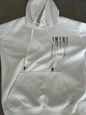 Vit hoodie från Amiri med tryck - Snygg vit hoodie från Amiri med stort färgglatt tryck på bröstet där bokstäverna har droppande effekt i svart, blått och orange. Hoodien har klassisk känguruficka och justerbar huva med snören. Perfekt för dig som gillar streetwear och unik design.