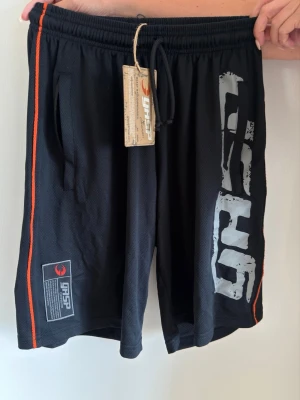 Svarta träningsshorts från GASP - Svarta träningsshorts från GASP med stora grå logotryck på framsidan och orange detaljer längs sidorna. Meshmaterial som är lätt och andas, elastisk midja med dragsko och fickor på sidorna. Perfekta för gym och sport. Helt nya med prislapp