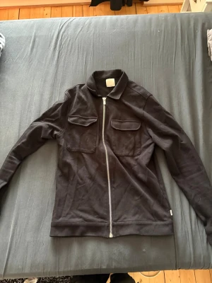 Svart overshirt med dragkedja - Säljer den då den inte passade mig. Den är i storlek L men är väldigt liten i storleken så skulle säga M/S. Svart overshirt med rak passform, dragkedja framtill och två bröstfickor med lock. Långa ärmar med knapp vid ärmslut och klassisk krage. Den är från Jack and Jones.