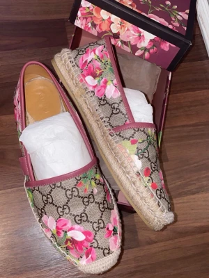 Gucci loafers med blommigt mönster - Säljer ett par loafers från Gucci med klassiskt GG-mönster och rosa blommor på beige canvas. Skorna har rosa läderdetaljer och flätad sula i jute. Perfekt för dig som vill sticka ut med snygga och trendiga skor.