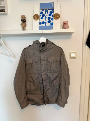 Grå vindjacka med huva - Snygg grå vindjacka med huva och dragkedja framtill. Jackan har två stora bröstfickor med lock, sidofickor och axelklaffar. Tillverkad i ett lätt, vindtätt material som passar perfekt för blåsiga dagar.
