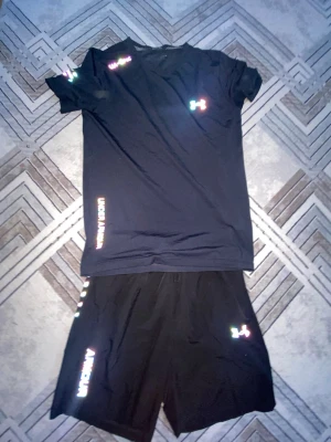 Svart träningsset från Under Armour - Snyggt svart träningsset från Under Armour med t-shirt och shorts. Båda plaggen har logga och textdetaljer i vitt. Materialet är lätt och andas, perfekt för träning. Shortsen har elastisk midja och t-shirten har en klassisk passform.