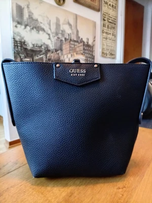 Svart bucket bag från Guess - Stilren svart bucket bag från Guess med strukturmönstrat konstläder och guldfärgade detaljer. Väskan har en rymlig insida och klassisk logga framtill. Perfekt för dig som vill ha en snygg och praktisk handväska med modern känsla.