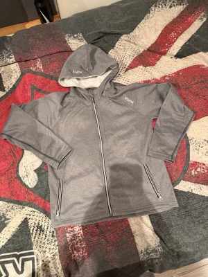Grå hoodie från Funk med dragkedja - Snygg grå hoodie från Funk med hel dragkedja framtill och två fickor med dragkedja. Hoodien har en mjuk insida och en klassisk huva. Perfekt för en avslappnad stil och enkel att matcha med andra plagg.