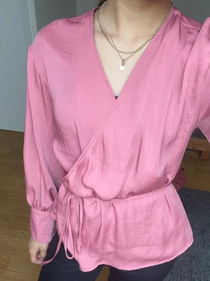 Rosa omlottblus med knytband - Trendig rosa omlottblus med långa ärmar och knytband i midjan. Blusen har en djup V-ringning och är tillverkad i ett mjukt, lätt material som ger ett snyggt fall. Perfekt för dig som vill ha en feminin och stilren look.