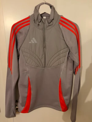 Grå träningsjacka från Adidas - Snygg grå träningsjacka från Adidas med röda detaljer och klassiska tre ränder på axlarna. Halvlång dragkedja framtill och hög krage. Tillverkad i ett stretchigt funktionsmaterial som passar perfekt till träning eller uppvärmning.