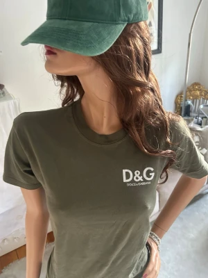Grön D&G t-shirt i bomull - Snygg mörkgrön topp från Dolce & Gabbana med vit D&G-logga på bröstet. Klassisk rund halsringning och korta ärmar. Tillverkad i mjuk bomull för en bekväm och avslappnad stil.